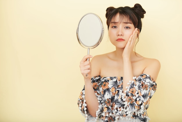 Young Asian woman holding up hand mirror and looking at camera with unhappy face