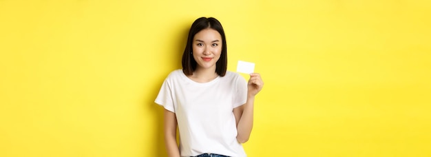 무료 사진 캐주얼 흰색 티셔츠에 플라스틱 신용 카드를 보여주고 카메라 노란색 바에서 웃는 젊은 아시아 여성