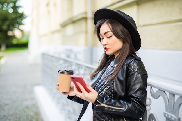 Giovane lettura asiatica della donna facendo uso delle notizie della lettura del telefono o degli sms mandanti un sms sullo smartphone mentre bevendo caffè sulla pausa da lavoro.