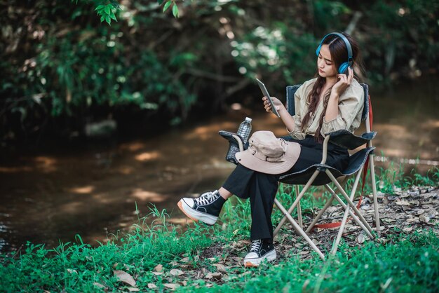 若いアジア人女性は、森の中でキャンプしながら、ワイヤレスヘッドフォンでタブレットから音楽を楽しんで聞いている小川の近くの椅子に座っています