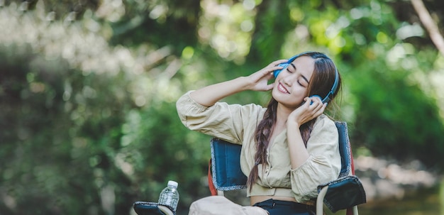 젊은 아시아 여성은 무선 헤드폰으로 음악을 들으며 시내 근처의 의자에 앉아 숲에서 캠핑을 하는 동안 행복하게 태블릿을 사용합니다.
