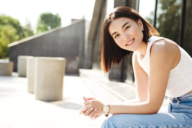 Young asian woman sitting on bench near building, smiling at camera with happy face