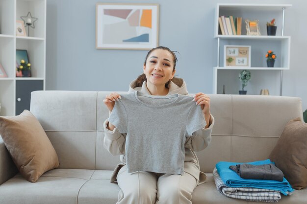 Young asian woman sitting on a couch at home interior holding stack of clean laundry happy and positive smiling