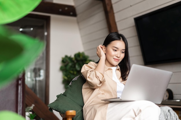 Молодая азиатская женщина, сидящая дома с девушкой портативного компьютера, просматривающая веб-сайты или обучающаяся удаленно