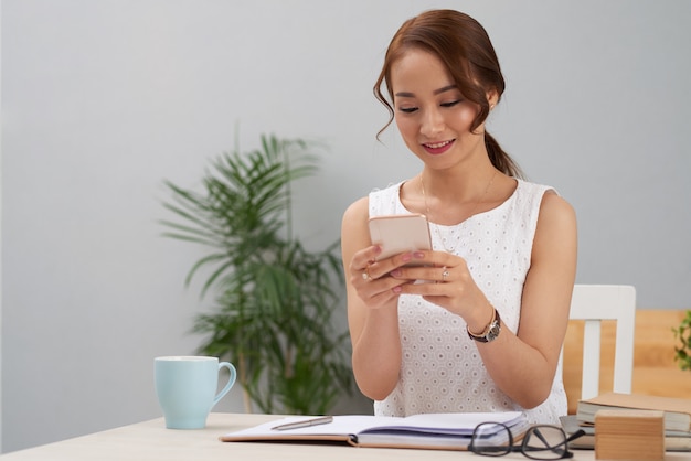 Young Asian woman sitting at table indoors and using smartphone