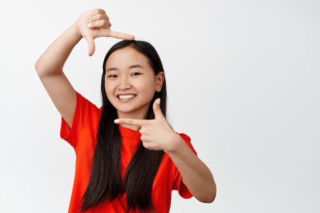 Young asian woman smiling, looking through hand frames, taking picture for happy memories, searching perspective, standing on white