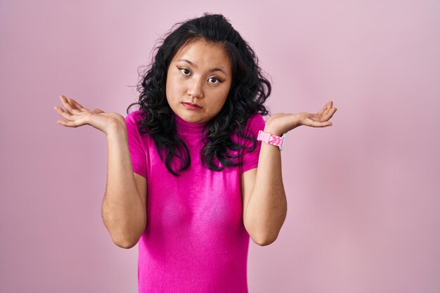 Young asian woman standing over pink background clueless and confused expression with arms and hands raised doubt concept