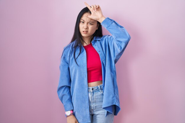 ピンクの背景の上に立っているアジアの若い女性は、額に指を置いて敗者のジェスチャーを嘲笑し、侮辱する人々をからかっています。