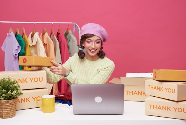 젊은 아시아 여성 스타트업 소규모 비즈니스 프리랜서 판매 패션 의류, 소포 상자와 컴퓨터 노트북이 탁자 위에 있고 분홍색 배경 온라인 마케팅 및 배달 개념에 격리되어 앉아 있습니다.