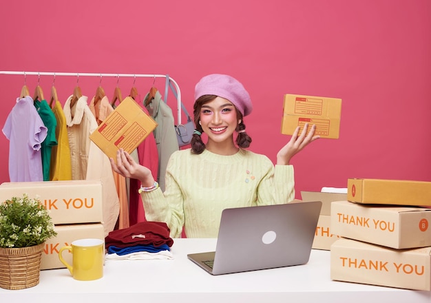 アジアの若い女性スタートアップ スモール ビジネス フリーランス販売ファッション衣料品小包ボックスとテーブルの上にコンピューターのラップトップとピンクの背景に分離されて座っているオンライン マーケティングと配信の概念