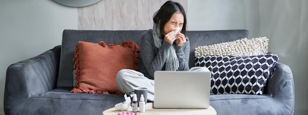 Young asian woman staying at home feeling unwell catching a cold sick leave sitting with laptop