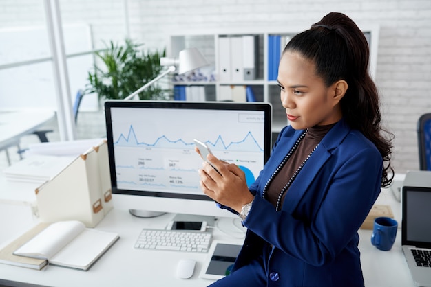 Free photo young asian woman in suit sitting on desk in office and using smartphone