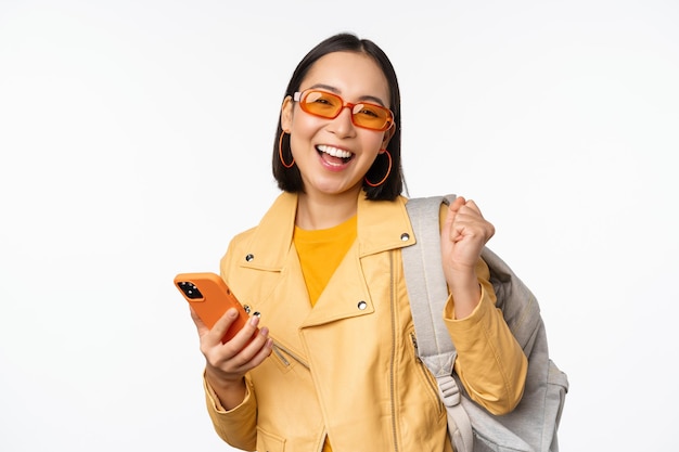 Young asian woman traveller girl tourist in sunglasses holding backpack and mobile phone using application on smartphone standing over white background