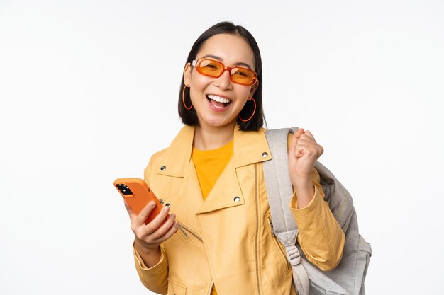 白い背景の上に立っているスマートフォンのアプリケーションを使用してバックパックと携帯電話を保持しているサングラスで若いアジアの女性旅行者の女の子の観光客