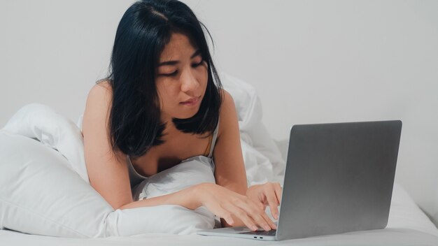 Young Asian woman using laptop checking social media feeling happy smiling while lying on bed after wake up at house in the morning, Attractive thai girl smiling relax in bedroom at home .