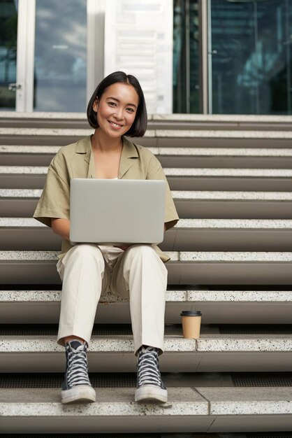 Молодая азиатская женщина, использующая ноутбук на открытом воздухе, милая студентка с компьютером делает домашнее задание, сидит