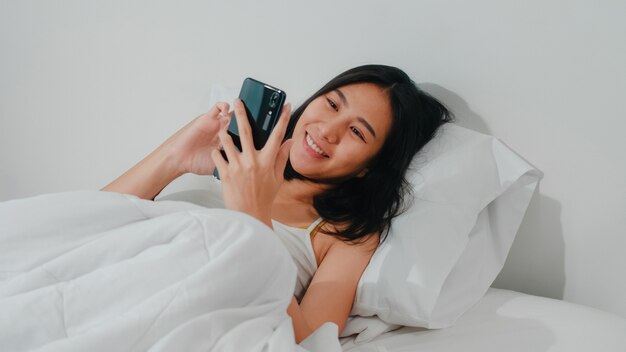 Молодая азиатская женщина используя smartphone проверяя социальные средства массовой информации чувствуя счастливый усмехаться пока лежащ на кровати после бодрствования в утре, красивый привлекательный усмехаться испанской дамы ослабляет в спальне дома.