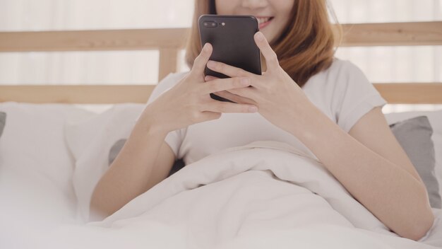 Молодая азиатская женщина используя smartphone лежа на кровати после встаю утром