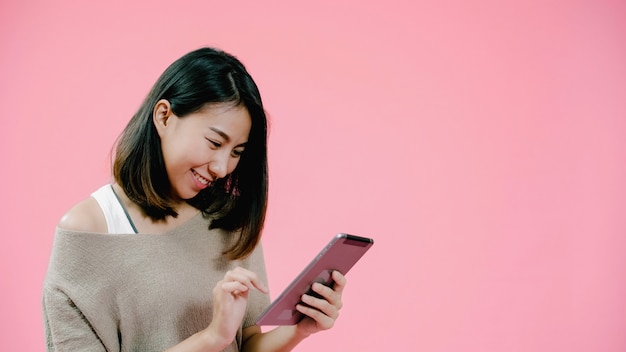 Young asian woman using tablet checking social media feeling happy smiling in casual clothing over pink background studio shot.