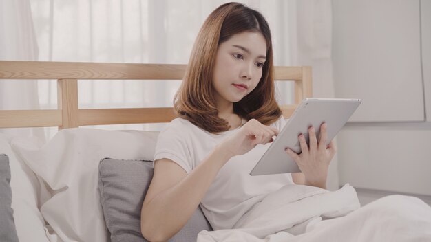 朝目を覚ます後ベッドに横たわっている間タブレットを使用して若いアジア女性