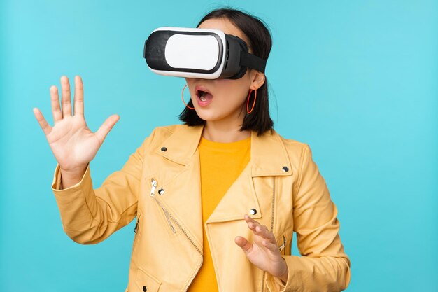 Молодая азиатка в очках виртуальной реальности с помощью гарнитуры VR, стоящая на синем фоне