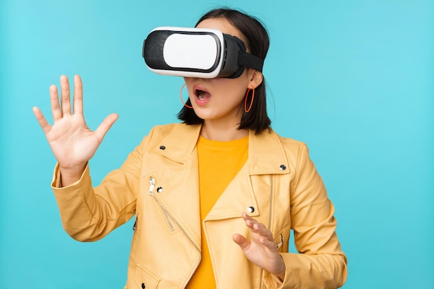 Young asian woman using virtual reality glasses using VR headset standing amused against blue background Copy space