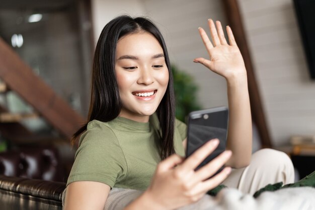 セルフォでチャットしているスマートフォンのカメラで手を振って携帯電話で若いアジアの女性のビデオ通話...