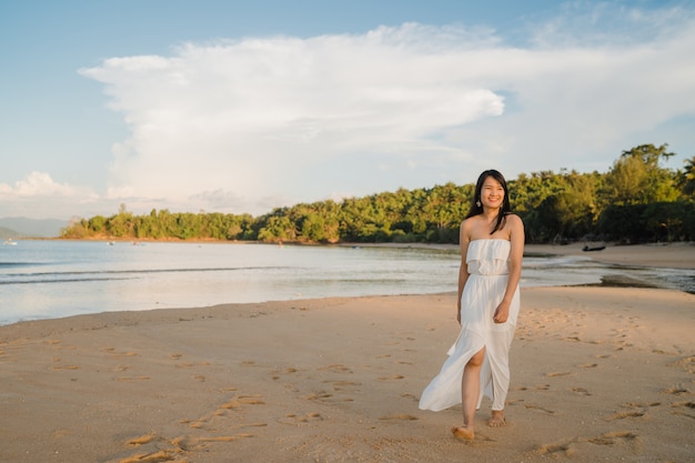 Молодая азиатская женщина гуляя на пляж. Красивое женское счастливое ослабляет идти на пляж около моря когда заход солнца в вечере.