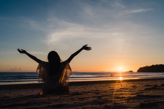 Молодая азиатская женщина наблюдая заход солнца около пляжа, красивое женское счастливое ослабляет наслаждается моментом когда заход солнца в вечере.