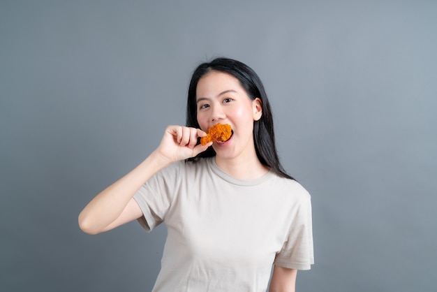 幸せそうな顔でtシャツを着て、灰色の壁にフライドチキンを食べることを楽しむ若いアジアの女性 プレミアム写真