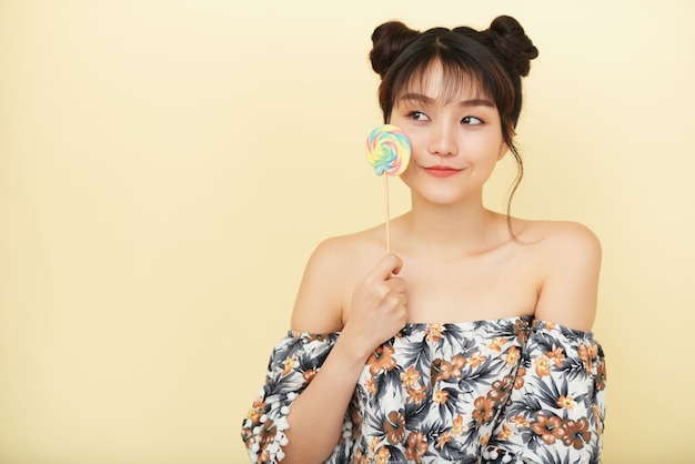 Young Asian woman with bare shoulders posing in studio and holding lollipop to cheek