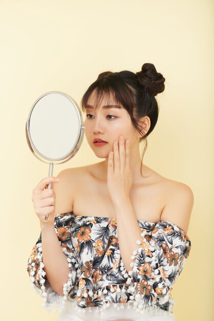 Young Asian woman with bare shoulders posing in studio and looking in hand mirror