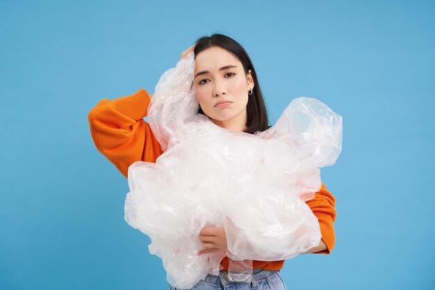 Young asian woman with plastic waste looking bothered by recycling standing over blue background