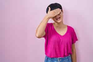 Бесплатное фото Молодая азиатская женщина с короткими волосами, стоящая на розовом фоне, улыбается и смеется, закрывая глаза рукой на лице для неожиданной слепой концепции