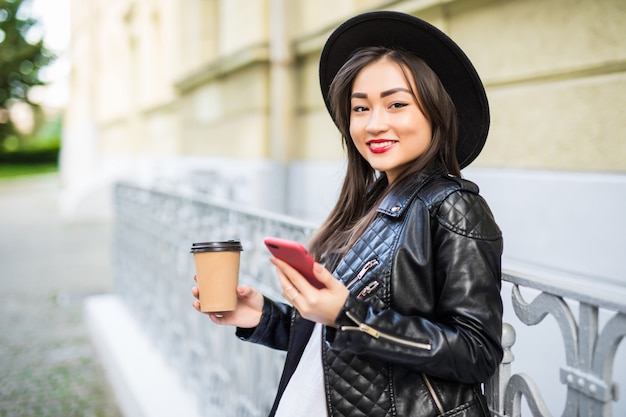 電話とコーヒーのカップが付いている通りに対してスマートフォンの立っている若いアジア女性