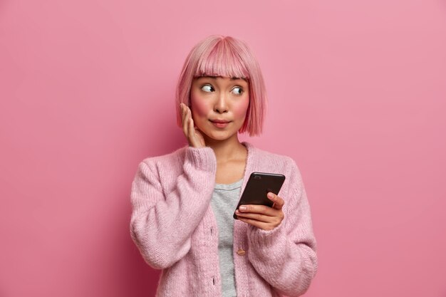 驚いた表情の若いアジア人女性は脇を向いて、カジュアルな服を着て、携帯電話を持って、ソーシャルメディアをサーフィンし、コンテンツ共有マルチメディアをオンラインで送信します。