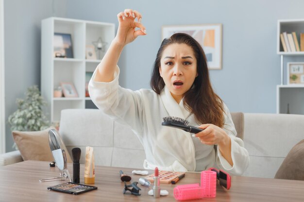 목욕 가운을 입은 젊은 아시아 여성이 가정 내 화장대에 앉아 머리를 빗고 아침 화장을 하는 동안 머리가 빠지는 것이 혼란스럽고 화가 났습니다.