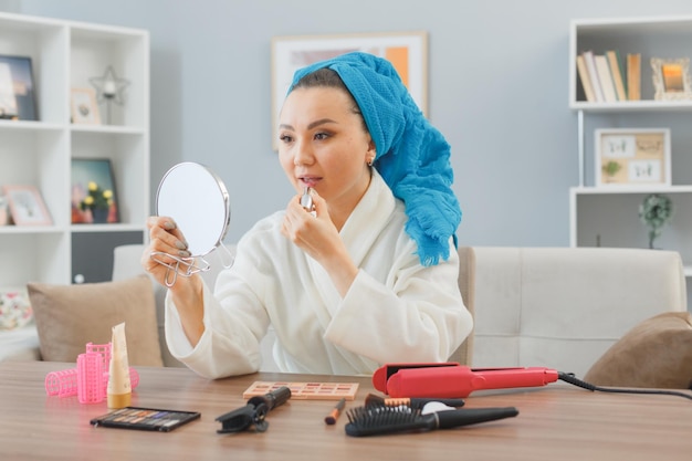 Молодая азиатка с полотенцем на голове сидит дома за туалетным столиком, наносит помаду и смотрит в зеркало, делая утренний макияж