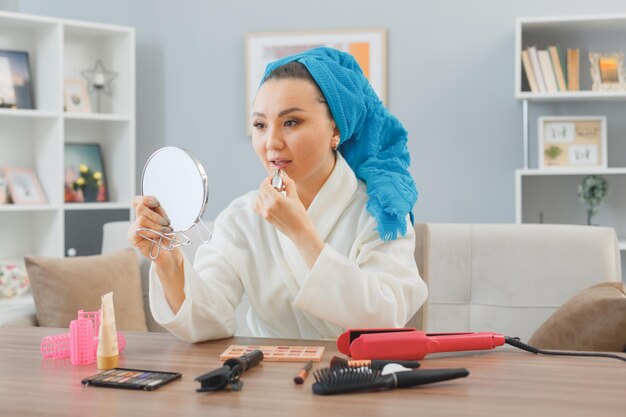 Young asian woman with towel on her head sitting at the dressing table at home applying lipstick looking in mirror doing morning makeup routine