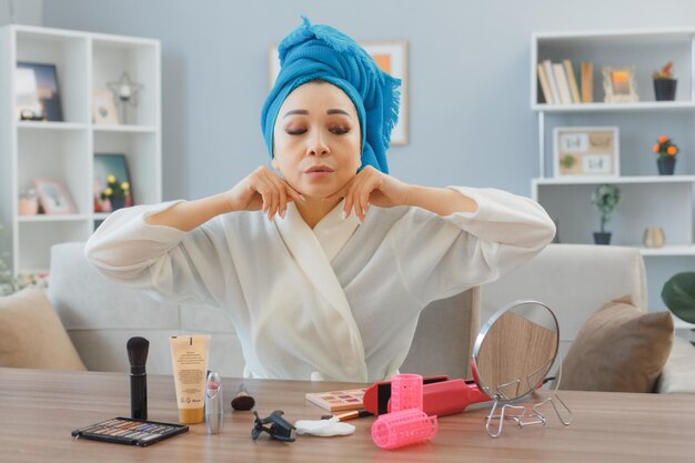 머리에 수건을 두른 젊은 아시아 여성이 집 내부 화장대에 앉아 아침 화장을 하는 일상적인 얼굴 마사지를 하고 있다