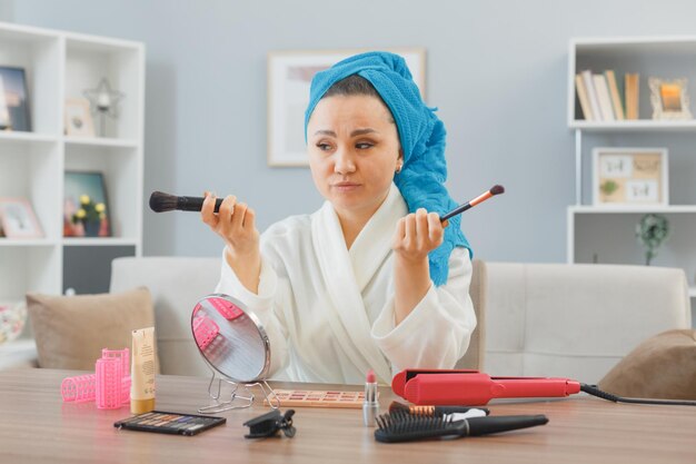 молодая азиатская женщина с полотенцем на голове сидит за туалетным столиком в домашнем интерьере, держа кисти для макияжа, смотрит в зеркало и смущенно делает утренний макияж