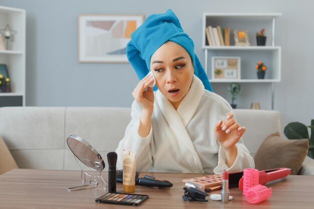 タオルを頭に抱えた若いアジア人女性が自宅の化粧台に座って鏡を見て、コットンパッドを顔に強壮剤を塗っている