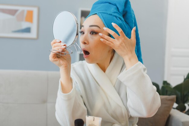 Молодая азиатская женщина с полотенцем на голове сидит за туалетным столиком в домашнем интерьере, смотрит в зеркало и смущается, касаясь глаз, делая утренний макияж