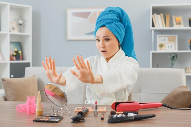 머리에 수건을 두른 젊은 아시아 여성이 집 화장대에 앉아 손톱이 아침 화장을 하는 것을 보며 화난 모습을 바라보고 있다