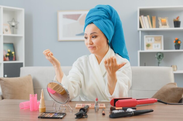 young asian woman with towel on her head sitting at the dressing table at home looking at her nails with skeptic expression doing morning makeup routine