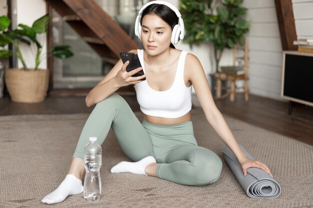 Young asian woman with water bottle listening music or podcast in headphones during fitness at home ...