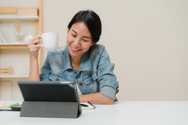 無料写真 タブレットを使用してソーシャルメディアをチェックしながらコーヒーを飲みながら働く若いアジア女性自宅のリビングルームの机の上。