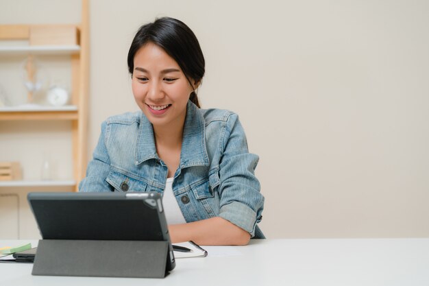 タブレットを使用してソーシャルメディアをチェックしながら働く若いアジア女性自宅のリビングルームの机の上。家のコンセプトで時間を楽しんでいます。