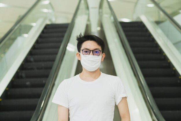 Young asiang glasses male wear protecting white mask to avoid corona virus or covid 19 spread asian man stand next to escalator urban lifestyle