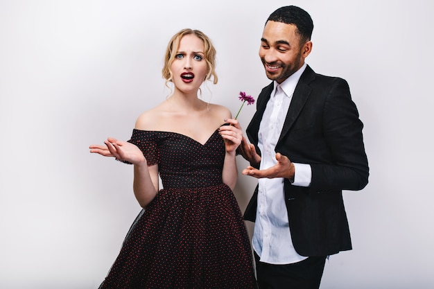 Free photo young astonished blonde woman in black luxury evening dress looking displeased with flower gift from handsome man in tuxedo smiling behind her. valentines day, present, having fun.